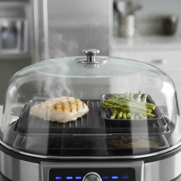 A close-up of a Power Smokeless Grill with a tempered glass lid, showing steam rising from vegetables on one side and grill marks forming on a steak on the other. The setting is a modern kitchen with a focus on healthy and efficient cooking.