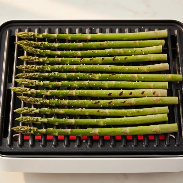 Seasoned asparagus spears arranged neatly on a hot electric grill surface, showing grill marks.